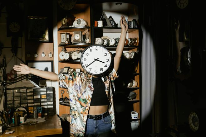 Someone with a clock for a face poses dramatically in a spotlight, shelves of clocks behind them.
