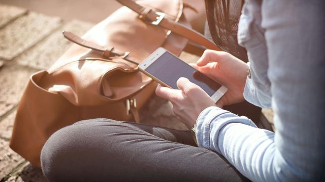 A woman sat crossed-legged on the ground with her bag, looking down at her smartphone.