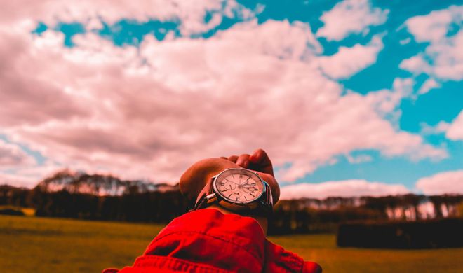 A closed hand reaches out from behind the camera, a watch on the wrist as the main focus, with a green expanse and blue sky in the background.