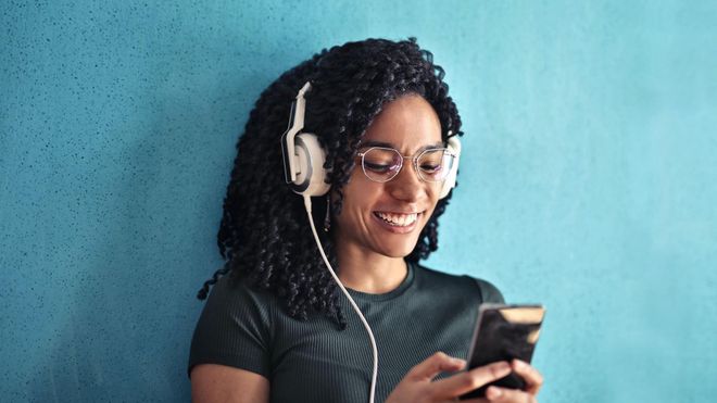 Woman of colour smiling while wearing glasses and large white headphones, looking at a mobile phone.