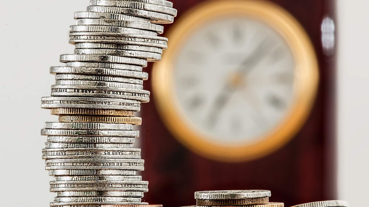 Close-up photo of a stack of coins with a wall clock out of focus in the background.