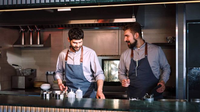 Two male kitchen staff wearing aprons talking while at work behind a counter.