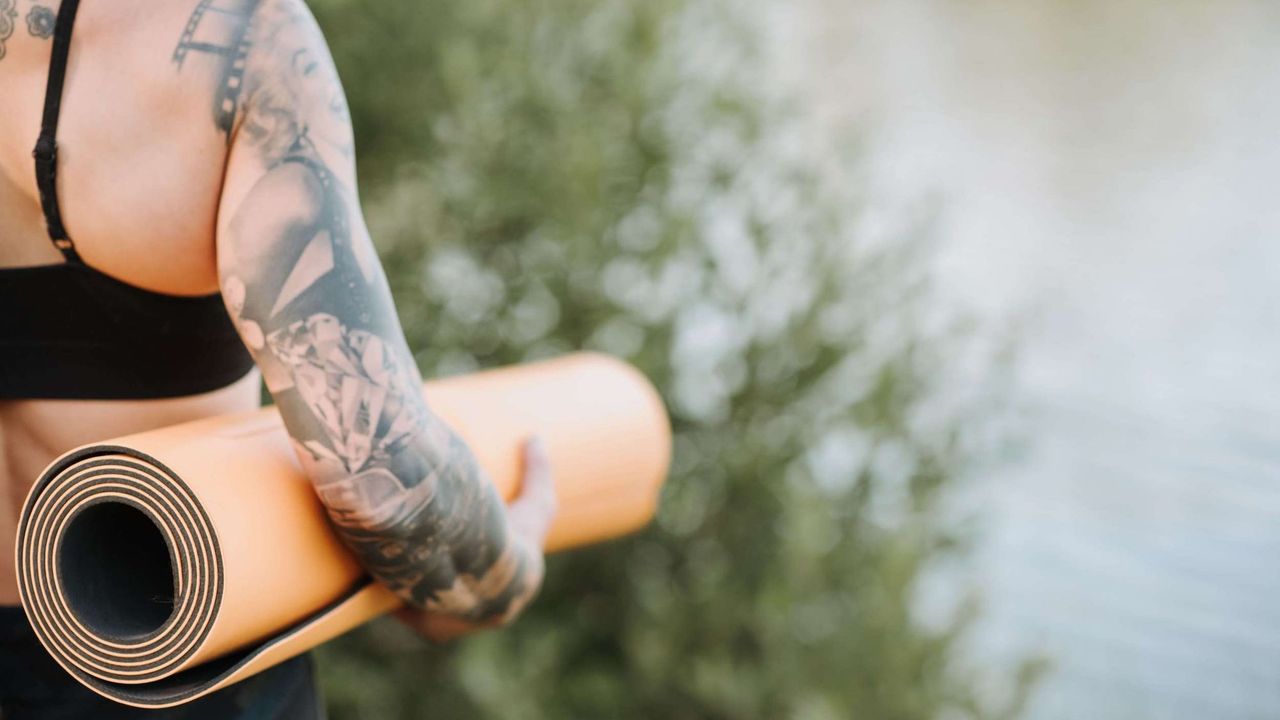 A tattooed woman holding an orange yoga mat under her arm, looking over a body of water.