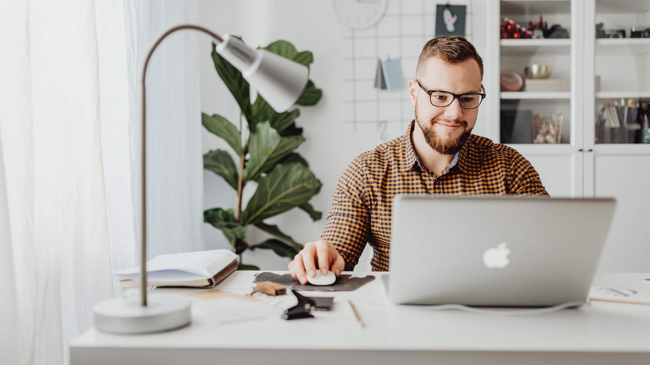 Photo of a man in checkered shirt smiling while using an Apple laptop at a desk