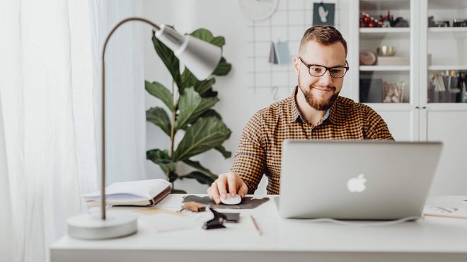 Photo of a man in checkered shirt smiling while using an Apple laptop at a desk
