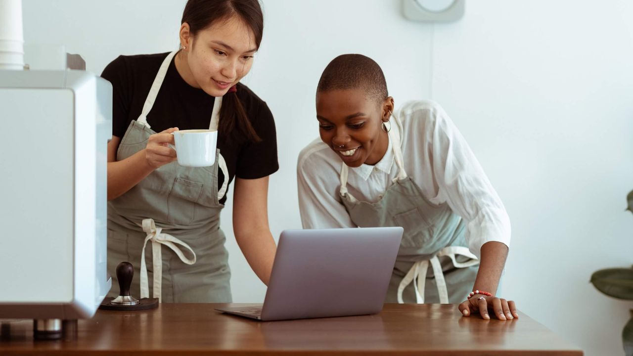 Two female baristas wearing aprons using a laptop computer on a wooden counter.