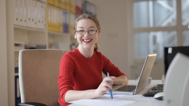 Woman wearing a red sweater smiling while working at a laptop