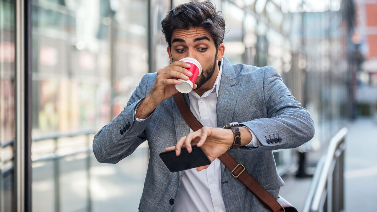 A suited man on his commute, looking at his watch in surprise as he drinks from a takeaway coffee cup.