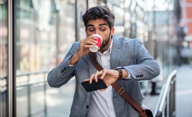 A suited man on his commute, looking at his watch in surprise as he drinks from a takeaway coffee cup.