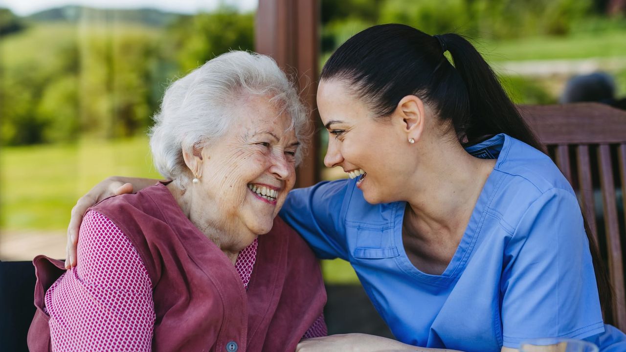 A carer has her arm around an elderly woman, both looking at each other and laughing.