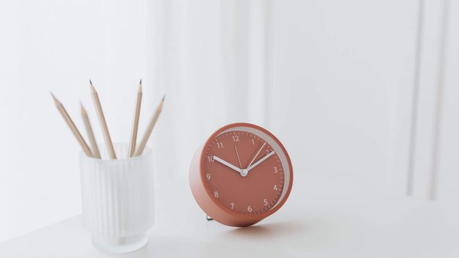 Photo of a pastel-pink clock on a white desk next to a cup filled with wooden pencils