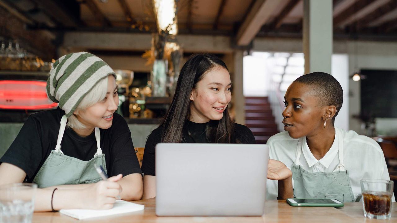 Three female baristas in conversation while sitting at a desk with a laptop computer.