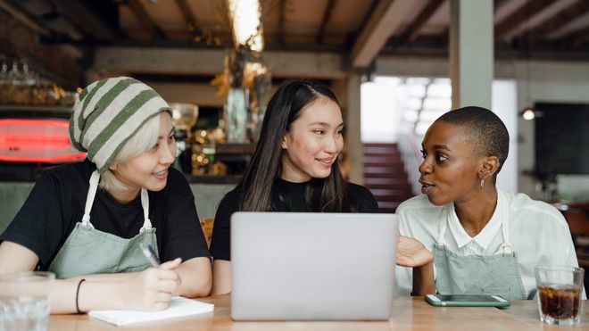 Three female baristas in conversation while sitting at a desk with a laptop computer.