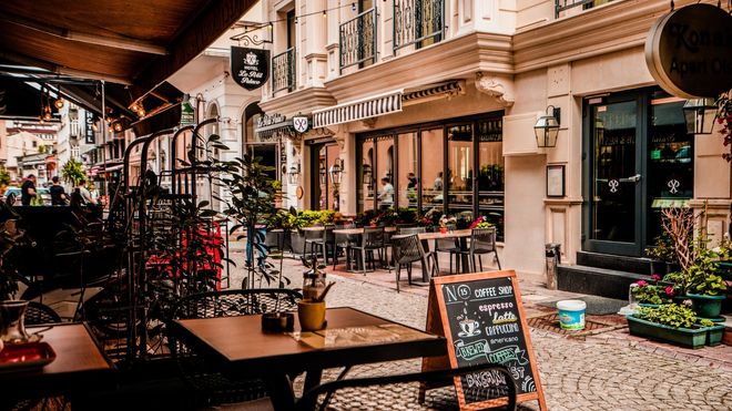 Cafe-lined cobbled street in a city centre, with awnings, outdoor tables and chairs, and a blackboard menu