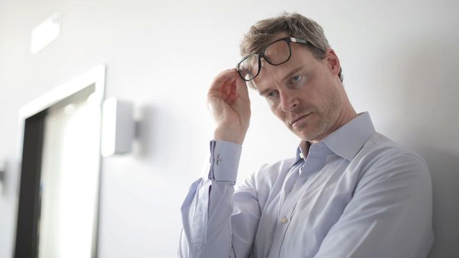 Annoyed-looking man in a shirt leaning against a wall, lifting his glasses off his face.
