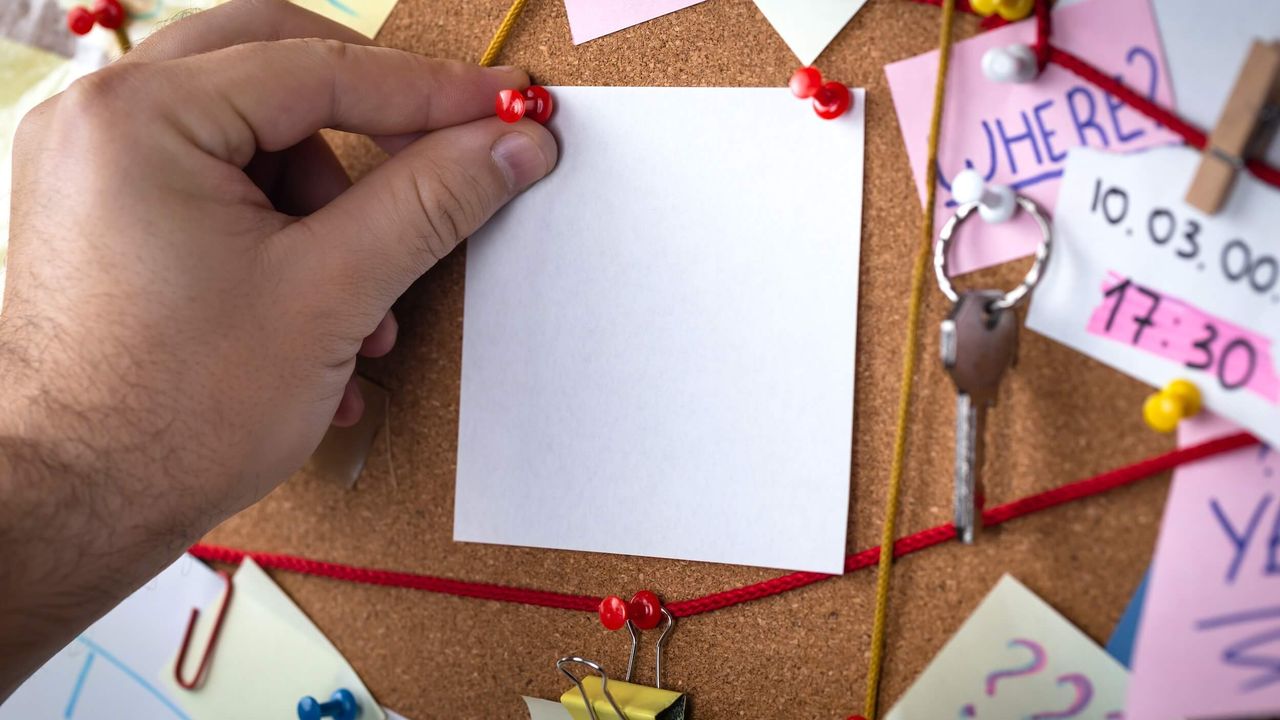 Close up photo of hand pinning a post-it note to a chaotic pinboard covered with notes