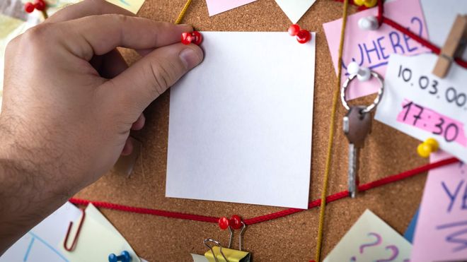 Close up photo of hand pinning a post-it note to a chaotic pinboard covered with notes