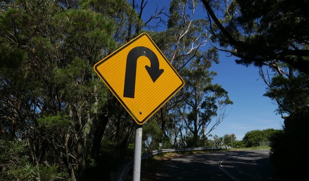 A yellow U-turn sign on a mountain road, flanked by trees and with a blue sky behind it.