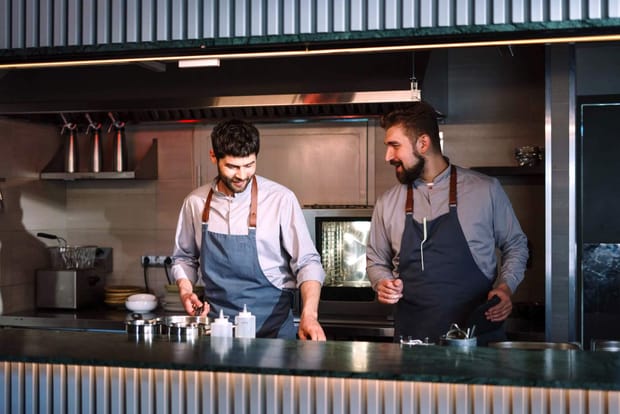 Two male kitchen staff wearing aprons talking while at work behind a counter.