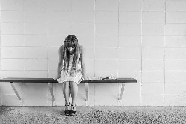 Woman with long hair sitting on a bench against a wall and looking down at her feet in black sandals