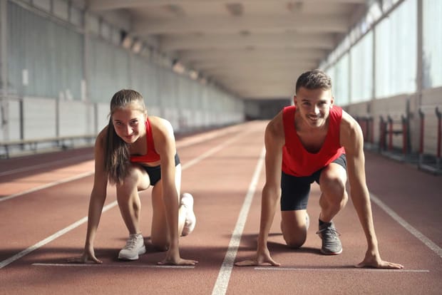 Female and male runner wearing red tank tops crouched on an indoor running track.