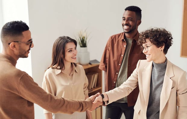 Woman with curly hair shaking hands with man wearing glasses as two other people look on, smiling.