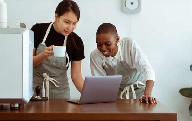 Two female baristas wearing aprons using a laptop computer on a wooden counter.