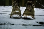 Close up of brown/green winter lace-up boots with snow on them, standing on snowy ground.
