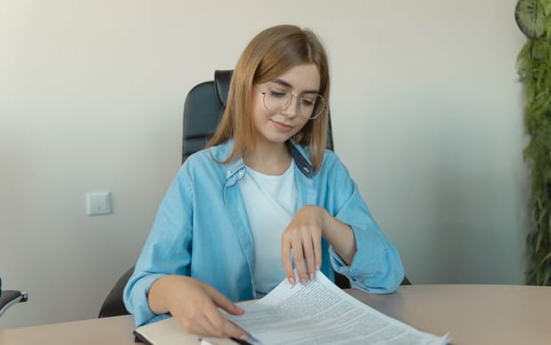 Woman wearing glasses and a light blue shift smiling while reading a paper document.