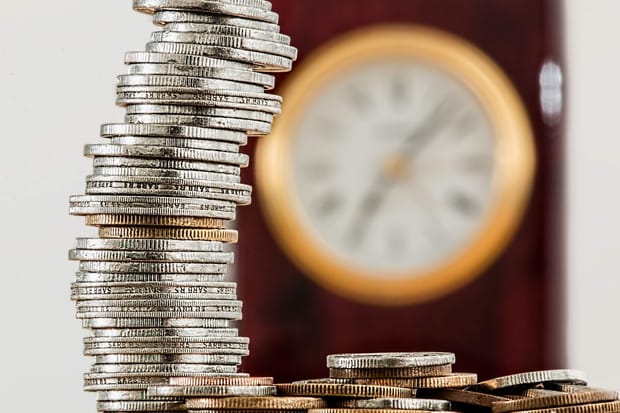 Close-up photo of a stack of coins with a wall clock out of focus in the background.