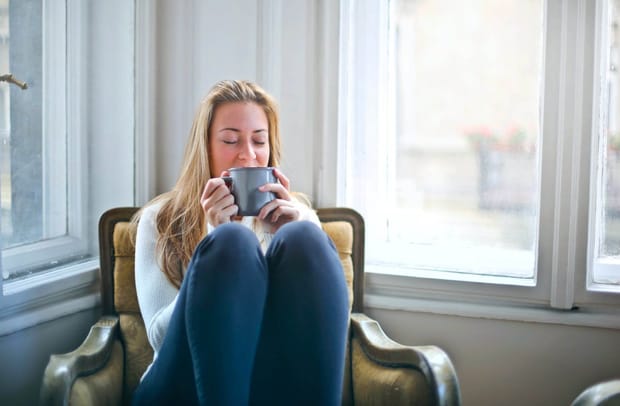Photo of a woman sitting in an armchair with her knees up, sipping from a ceramic mug