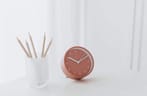 Photo of a pastel-pink clock on a white desk next to a cup filled with wooden pencils