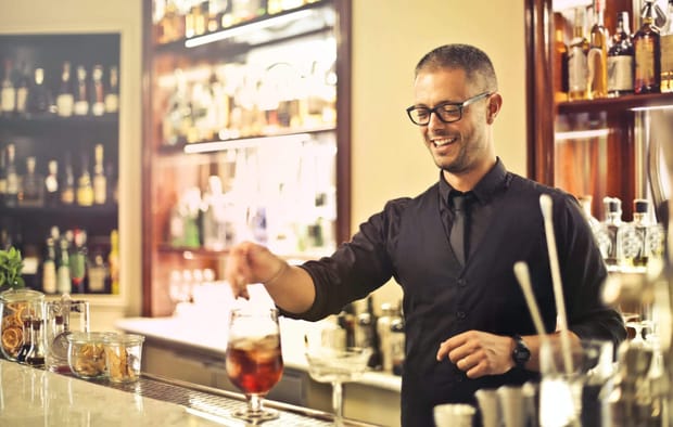 Photo of a bartender wearing a waistcoat and glasses smiling while preparing a cocktail on the bar