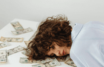 A brunette woman resting her head, tired, on a table that's covered in dollar bills.