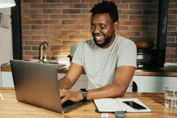 A young man sat at a kitchen counter with an open notebook and a laptop, smiling at the screen as he types.