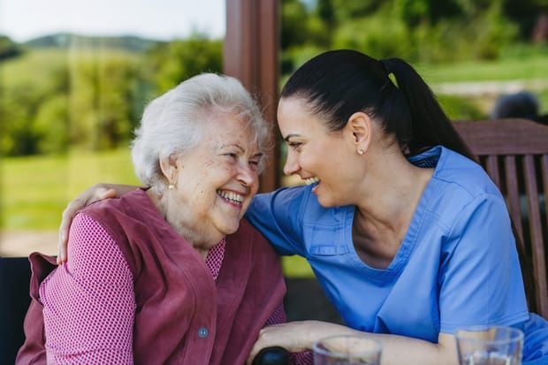 A carer has her arm around an elderly woman, both looking at each other and laughing.
