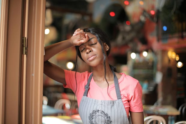A young female hospitality worker, leaning against a doorframe with her eyes closed and forehead pressed against the back of her hand.
