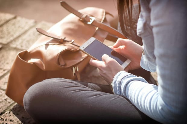 A woman sat crossed-legged on the ground with her bag, looking down at her smartphone.