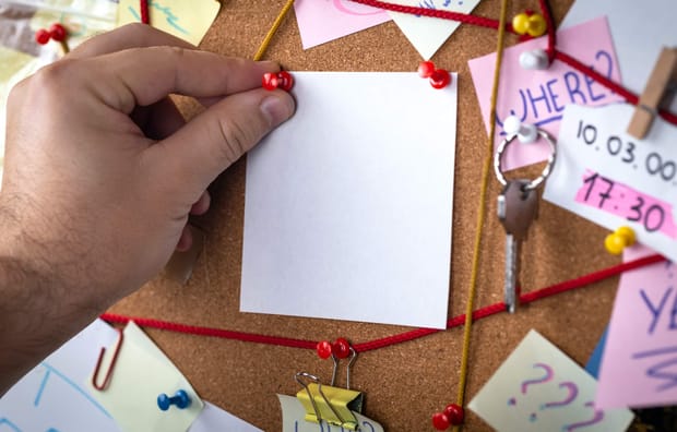 Close up photo of hand pinning a post-it note to a chaotic pinboard covered with notes
